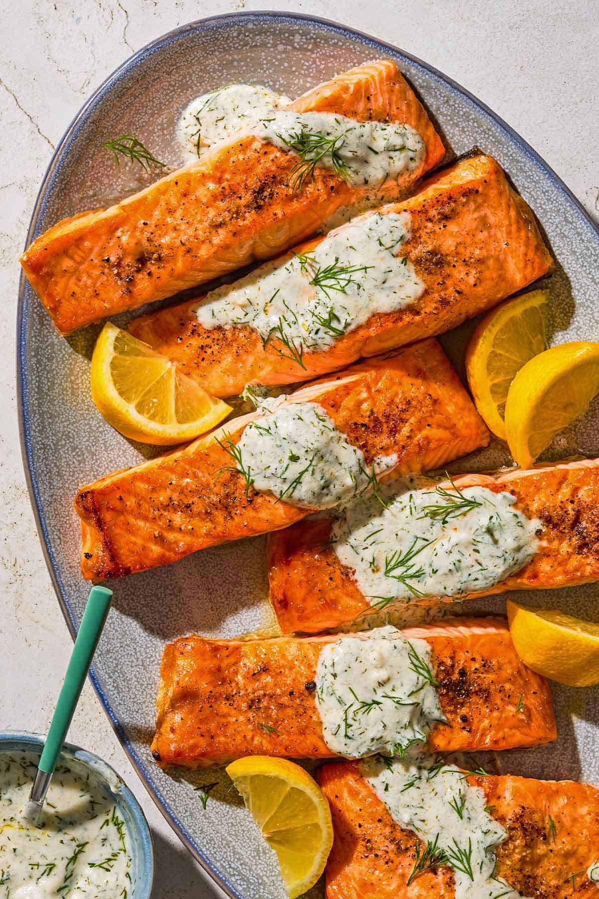 Six lemon dill salmon fillets on a platter with lemon wedges. Next to this is a bowl of dill sauce for salmon with a spoon.