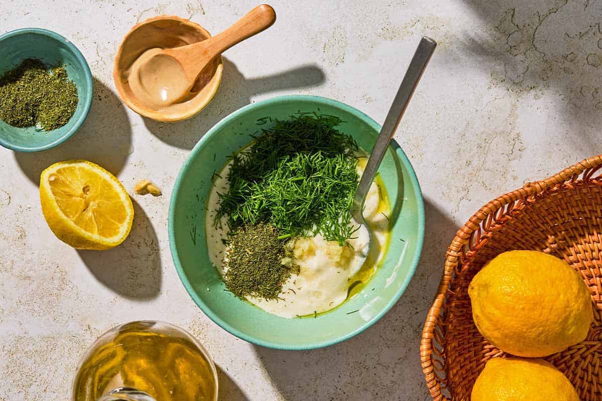 The ingredients for the dill sauce for salmon in a bowl with a spoon just before being mixed together. Next to this is a basket of lemons, a bottle of olive oil, a juiced lemon half, a bowl of dried dill, and a bowl of dijon mustard.