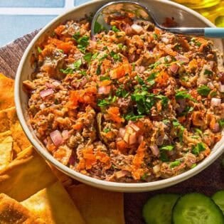 Sardine dip in a bowl with a spoon next to cucumber slices, pita chips, a bowl of olives and a bottle of olive oil.