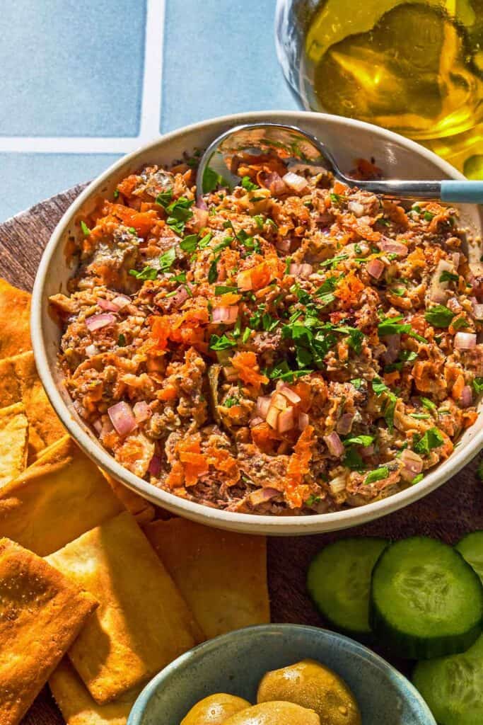 Sardine dip in a bowl with a spoon next to cucumber slices, pita chips, a bowl of olives and a bottle of olive oil.