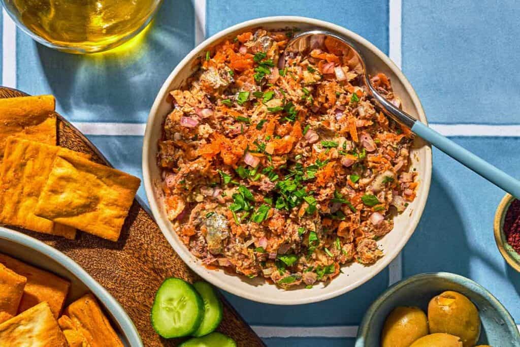 Sardine dip in a bowl with a spoon. Next to this is a platter with pita chips and sliced cucumbers, a bowl of green olives, a bowl of sumac and a bottle of olive oil.