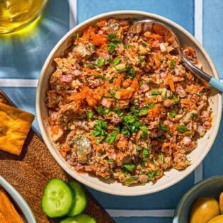 Sardine dip in a bowl with a spoon. Next to this is a platter with pita chips and sliced cucumbers, a bowl of green olives, a bowl of sumac and a bottle of olive oil.