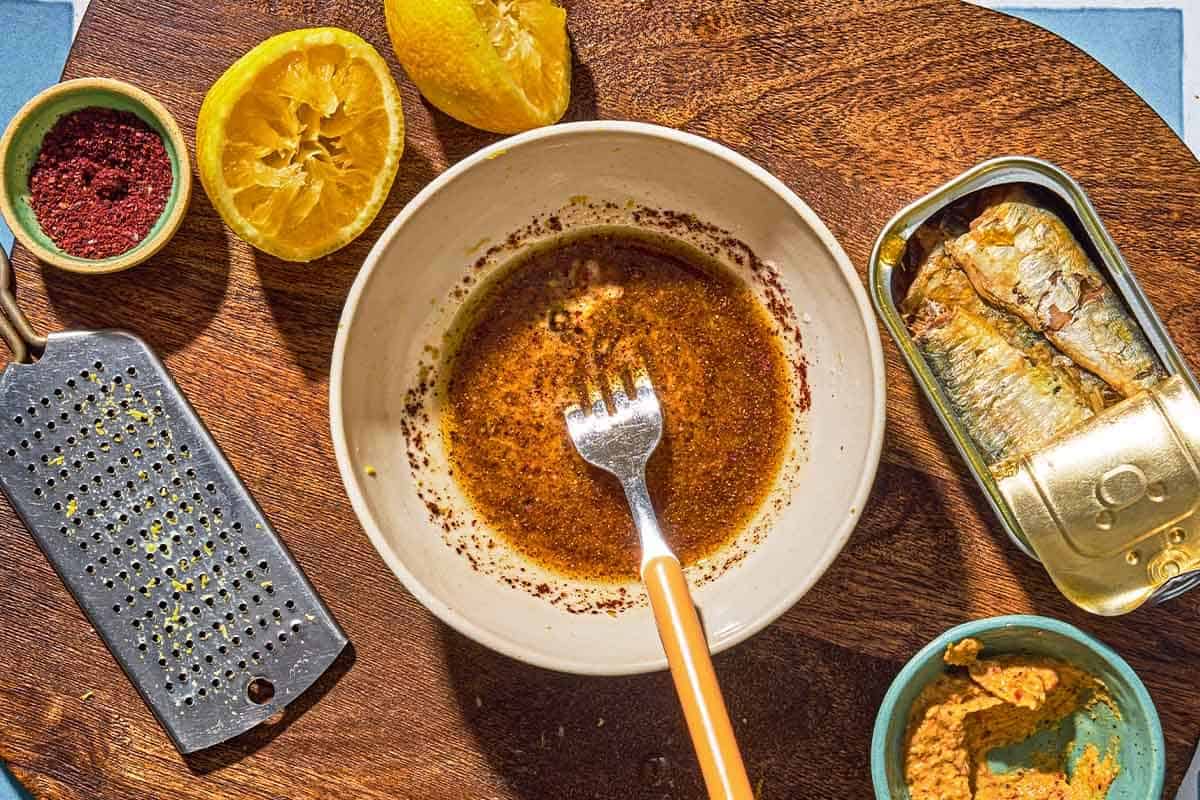 The dressing for the sardine dip in a bowl with a fork. Next to this is a zester, a bowl of sumac, 2 juiced lemon halves, a tin of sardines and a bowl of dijon mustard.