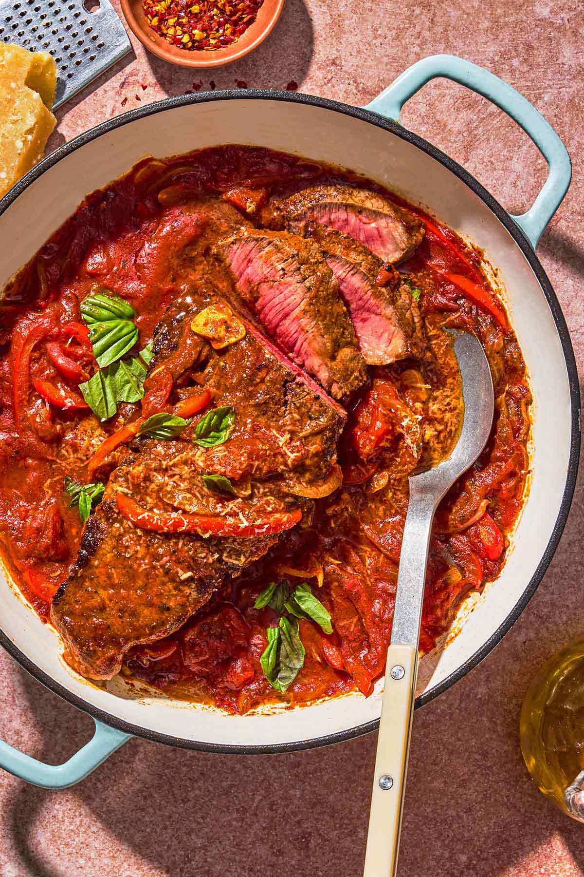 The steak pizzaiola in a skillet with a serving spoon. Next to this is a bowl of red pepper flakes, parmesan cheese and a cheese grater.