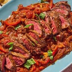 The steak pizzaiola on a platter next to parmesan cheese, a cheese grater and a bowl of red pepper flakes.