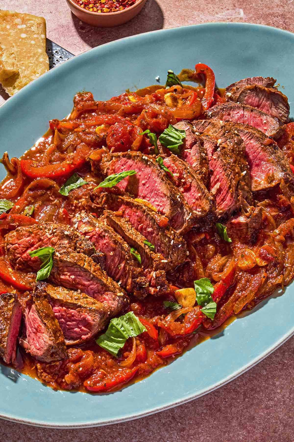 The steak pizzaiola on a platter next to parmesan cheese, a cheese grater and a bowl of red pepper flakes.