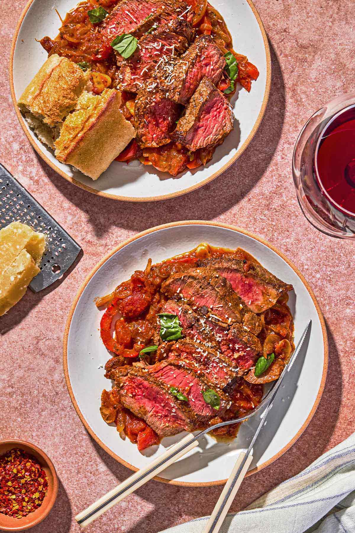 A serving of steak pizzaiola on a plate with a knife and fork. Next to this is a glass of red wine, another plate of the pizzaiola with some crusty bread, parmesan cheese, a cheese grater and a bowl of red pepper flakes.