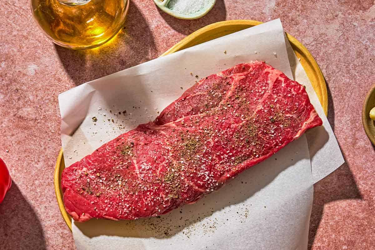 The uncooked seasoned steak for the steak pizzaiola on a parchment lined plate. Next to this is a bottle of olive oil and a bowl of salt.