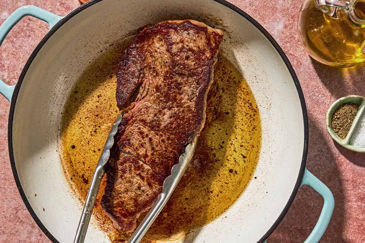 The steak for the steak pizzaiola being seared in a skillet. Next to this is a bottle of olive oil and a bowl of salt and pepper.