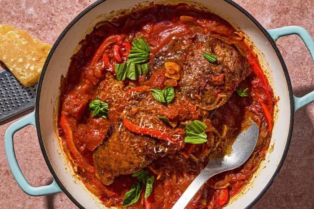 The steak pizzaiola in a skillet with a serving spoon next to parmesan cheese and a cheese grater.
