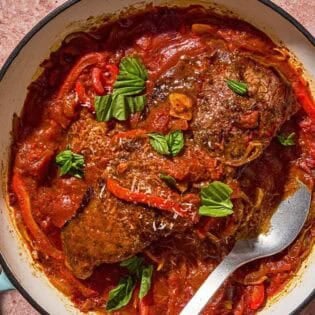 The steak pizzaiola in a skillet with a serving spoon next to parmesan cheese and a cheese grater.