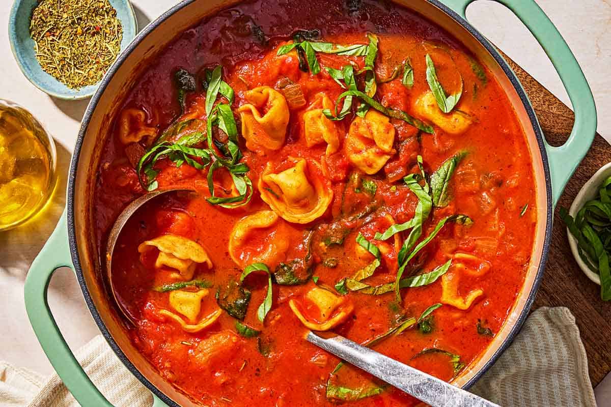 Tomato tortellini soup in a pot with a ladle. Next to this is a bottle of olive oil, and a bowl of spices.