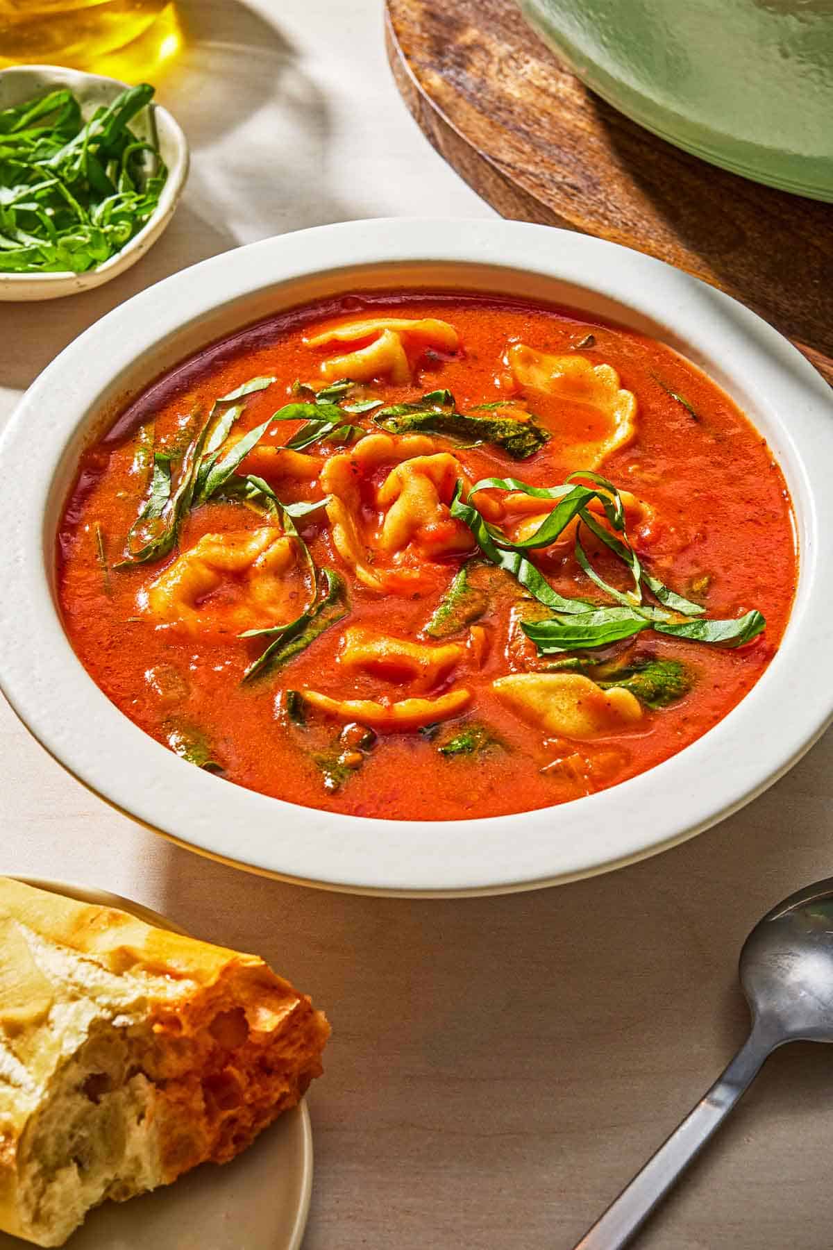 Tomato tortellini soup in a bowl next to a spoon, some crusty bread and a bowl of basil.