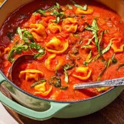 Tomato tortellini soup in a pot with a ladle. Next to this is a bottle of olive oil, and a bowl of spices.