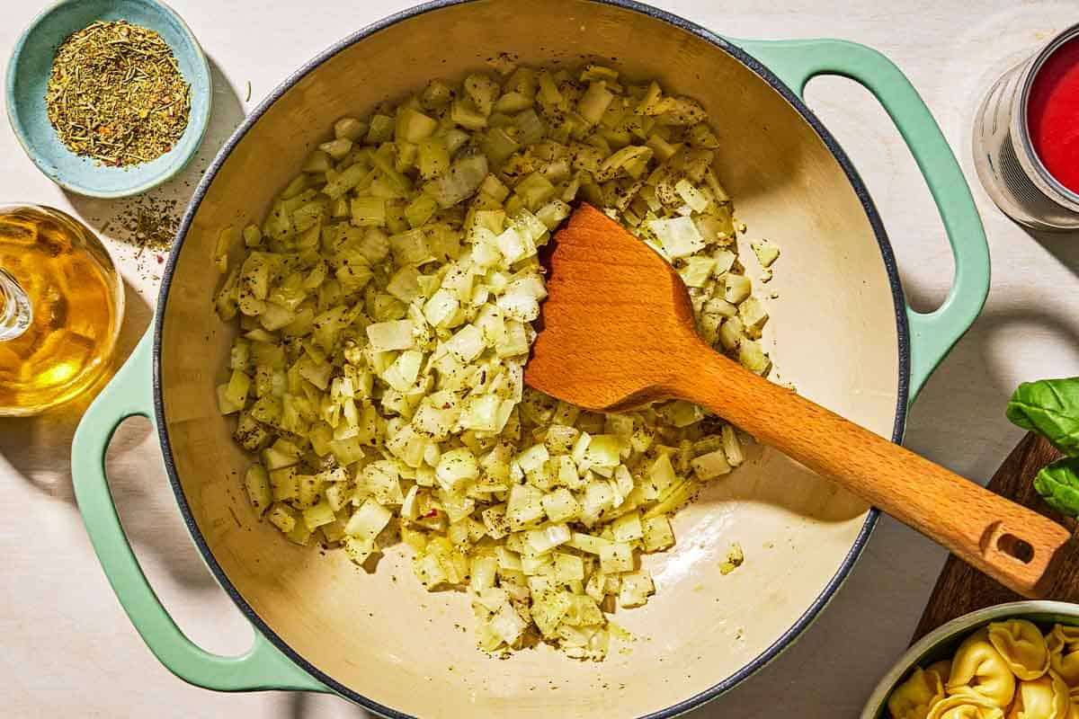 The aromatics for the tomato tortellini soup being sautéed in a pot with a wooden spatula. Next to this is a bottle of olive oil, a bowl of spices, a can for tomato sauce, and a bowl of cheese tortellini.