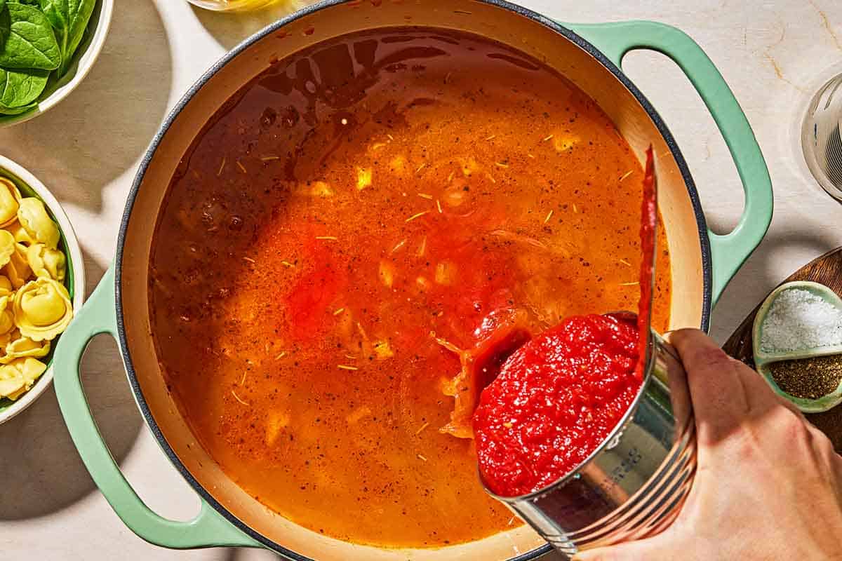 The crushed tomatoes being added to the tomato tortellini soup in a pot. Next to this are bowls of cheese tortellini, basil, salt and pepper.