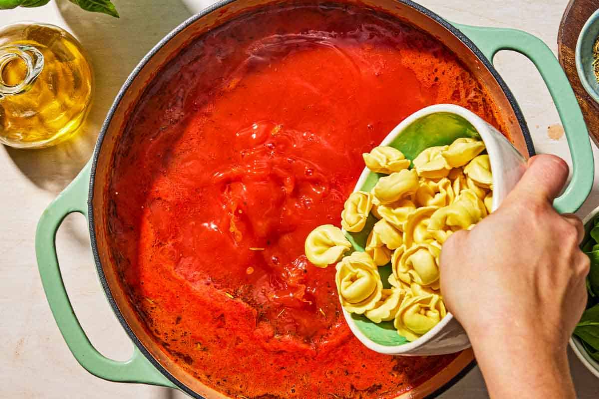 The cheese tortellini being added to the tomato tortellini soup in a pot. Next to this is a bottle of olive oil.
