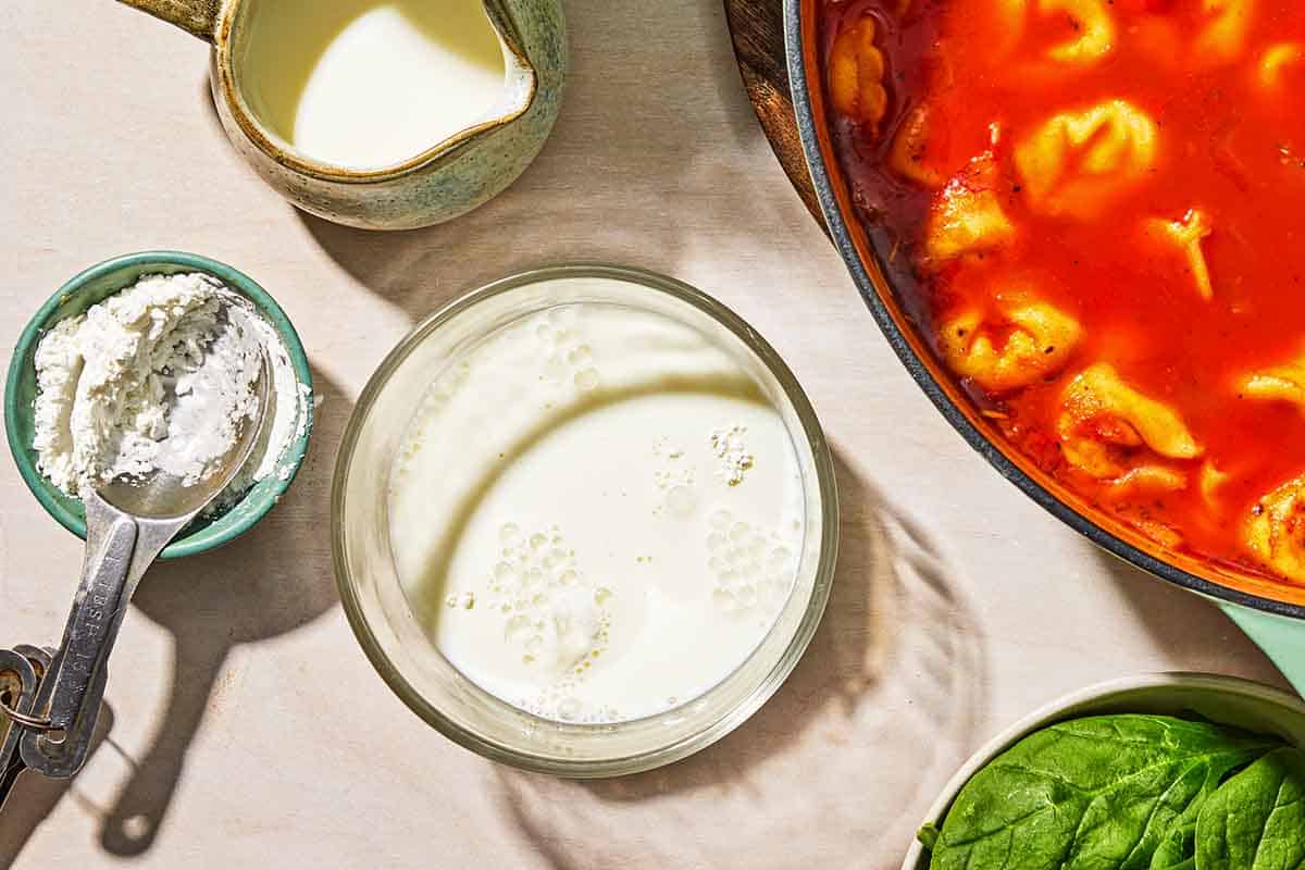 A bowl of the milk and cornstarch mixture for the tomato tortellini soup next to a bowl of cornstarch, a small pitcher of milk, a bowl of basil, and the soup in a pot.