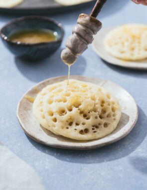 Honey being drizzled on 2 baghrir on a plate.