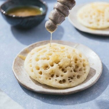 Honey being drizzled on 2 baghrir on a plate.