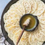 Several baghrir drizzled with honey surrounding a bowl of the honey and butter mixture with a honey dipper on a platter.