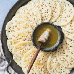 Several baghrir drizzled with honey surrounding a bowl of the honey and butter mixture with a honey dipper on a platter.