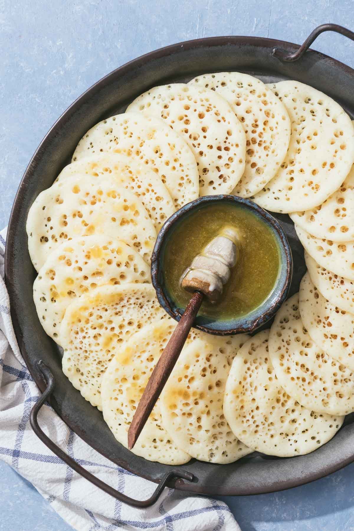 Several baghrir drizzled with honey surrounding a bowl of the honey and butter mixture with a honey dipper on a platter.