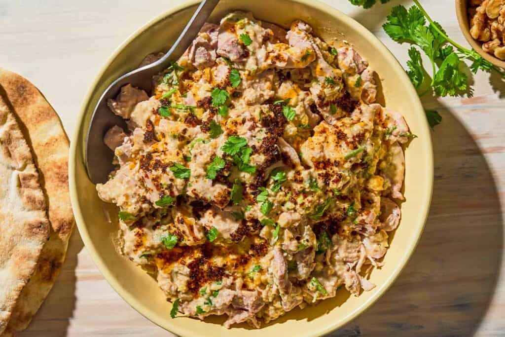 Circassian chicken in a bowl with a spoon next to 2 pieces of pita, sprigs of cilantro and a bowl of walnuts.