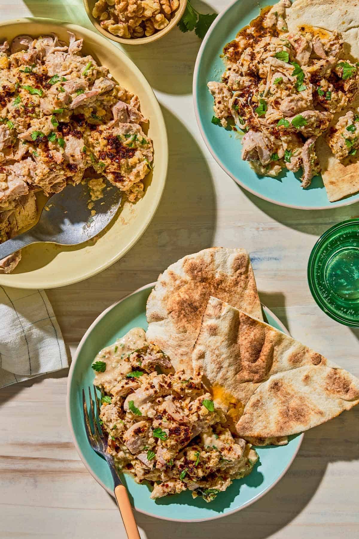 Two plates of the circassian chicken with pieces of pita. Next to this is the rest of the circassian chicken in a bowl with a spoon, a glass of sparkling water and a bowl of walnuts.
