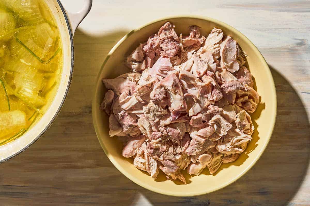 A bowl of shredded chicken next to the poaching liquid for the chicken in a pot.