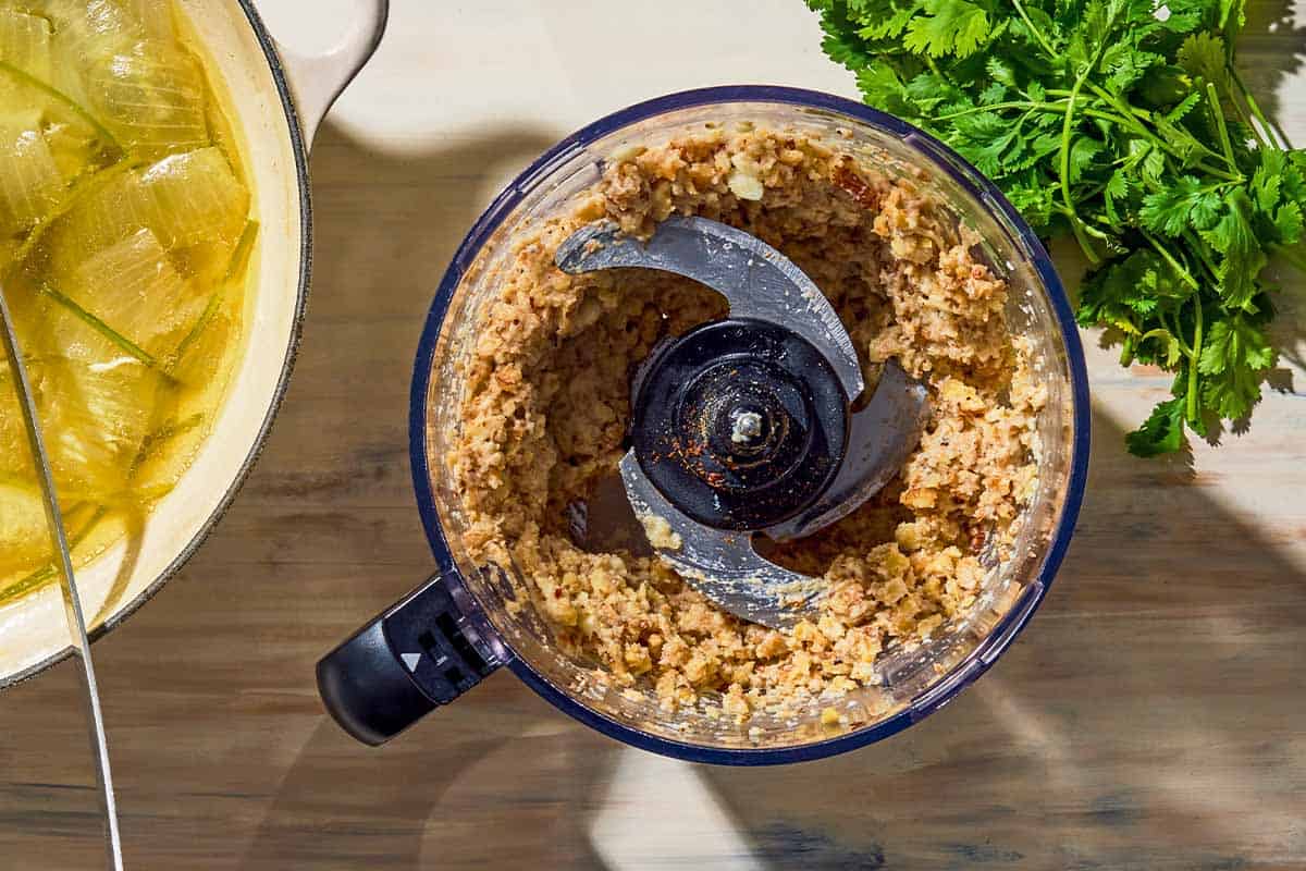 The ingredients for the circassian chicken mixed together in the bowl of a food processor fitted with a blade. Next to this is a bunch of cilantro and the poaching liquid for the chicken in a pot.