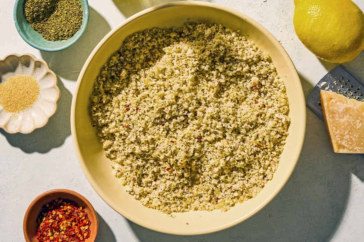 the oreganata for the clams oreganata in a bowl. Next to this is a wedge of parmesan cheese, a cheese grater, a lemon, and bowls of dried oregano, garlic powder and red pepper.