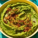 Fava bean dip topped with caramelized onions in a bowl next to a bowl of chili flakes and a bottle of olive oil.
