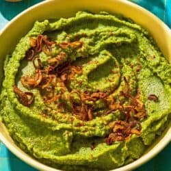 Fava bean dip topped with caramelized onions in a bowl next to a bowl of chili flakes and a bottle of olive oil.