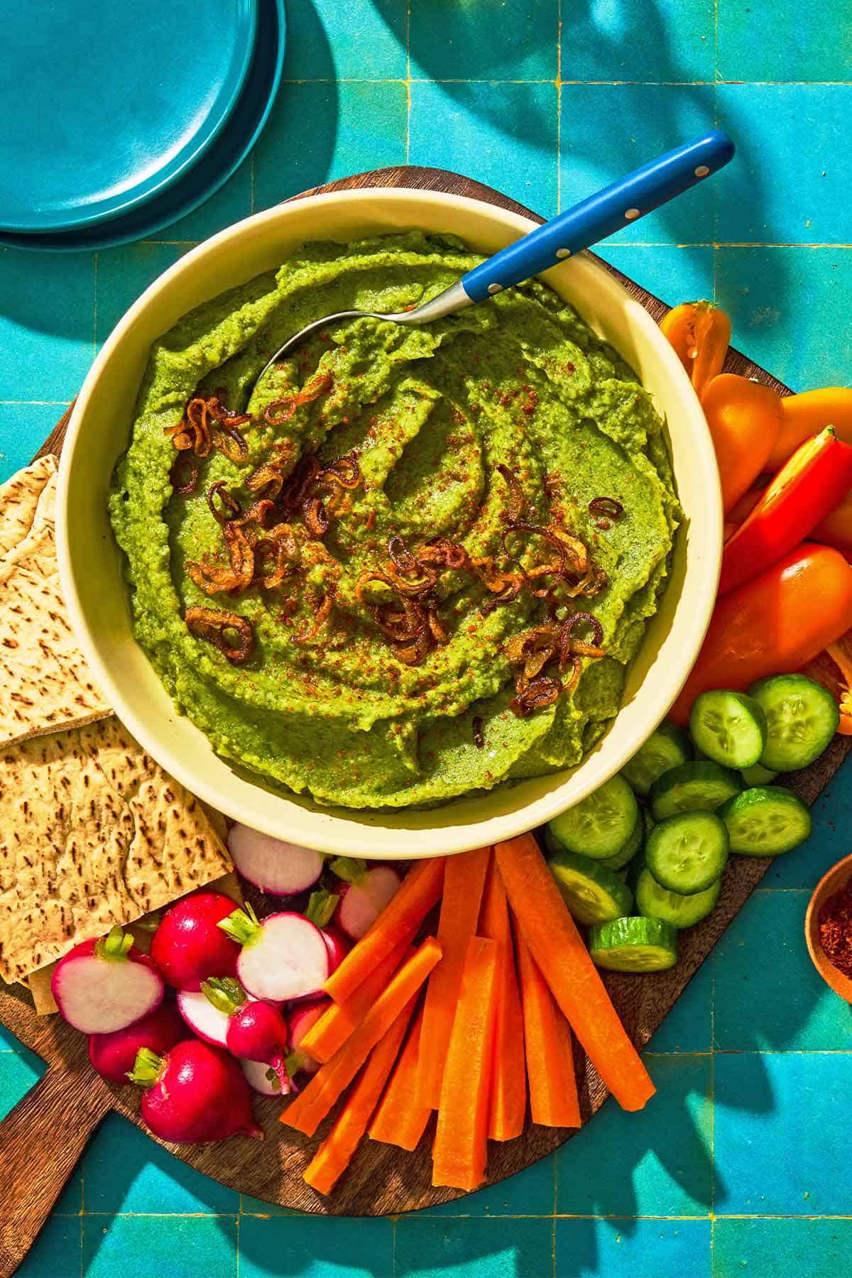 Fava bean dip topped with caramelized onions in a bowl on a wooden tray with various vegetables and more pita wedges. Next to this is a stack of two plates.