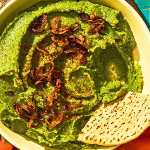 Fava bean dip topped with caramelized onions in a bowl with a pita wedge. This is on a platter with various vegetables and more pita wedges.