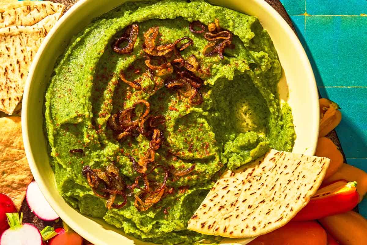 Fava bean dip topped with caramelized onions in a bowl with a pita wedge. This is on a platter with various vegetables and more pita wedges.