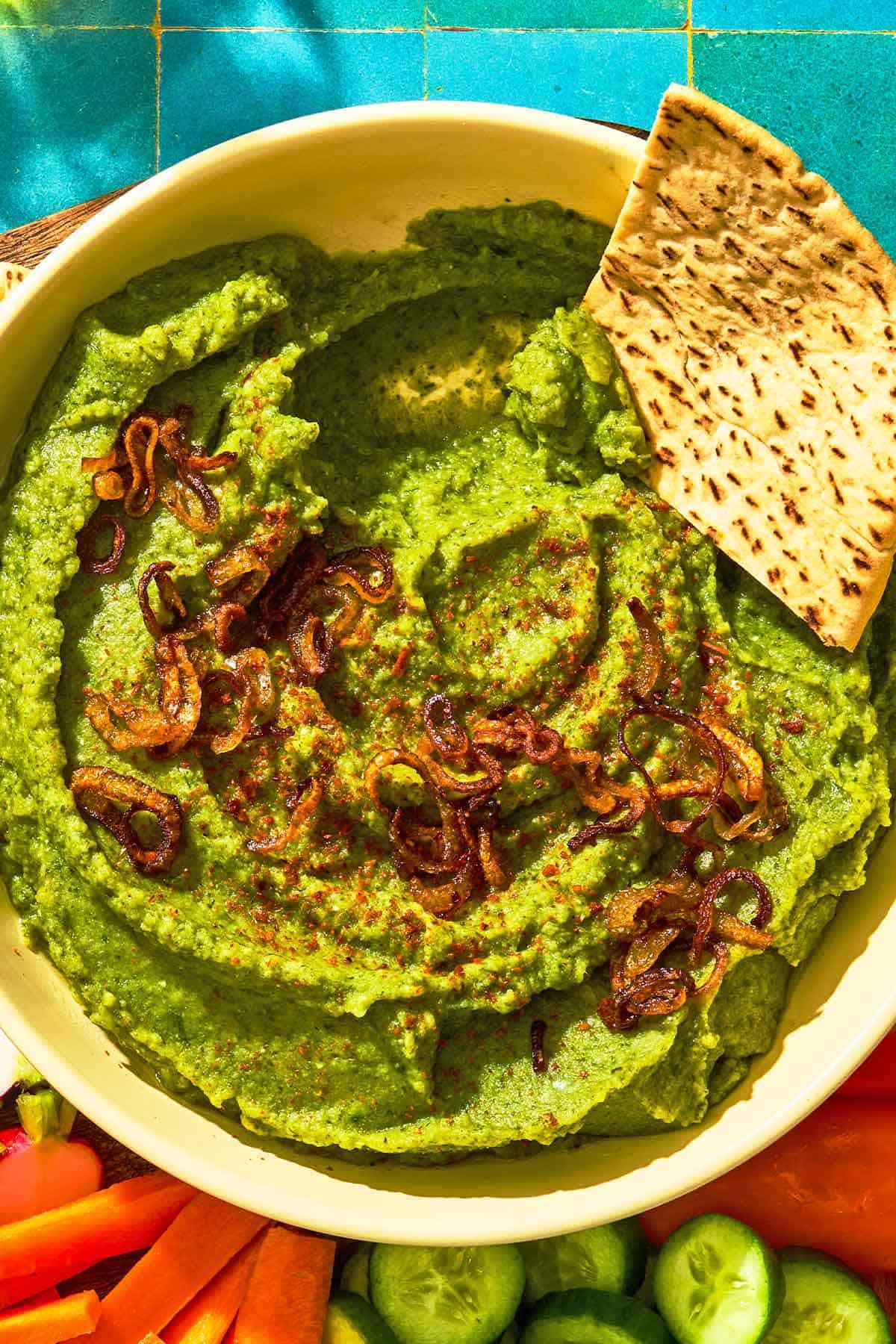 Fava bean dip topped with caramelized onions in a bowl with a pita wedge. This is on a tray with various vegetables.