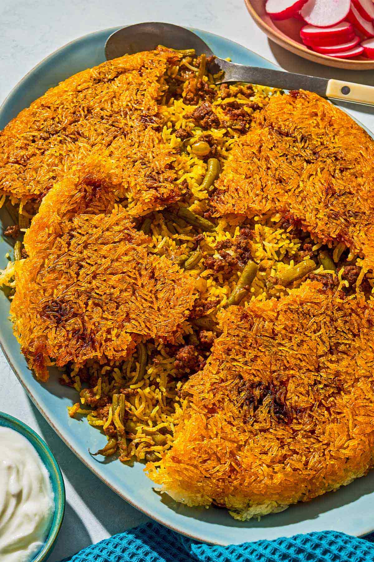 Loobia polo on a platter with a serving spoon. Next to this is a bowl of sliced radishes and a bowl of yogurt.