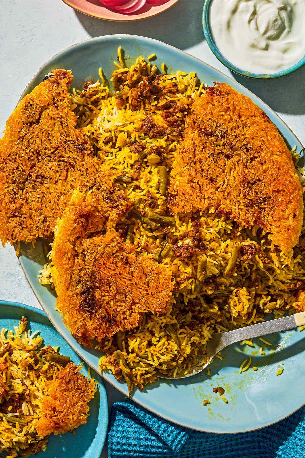 Loobia polo on a platter with a serving spoon. Next to this is a bowl of sliced radishes, a bowl of yogurt and a serving of the loobia polo on a plate.