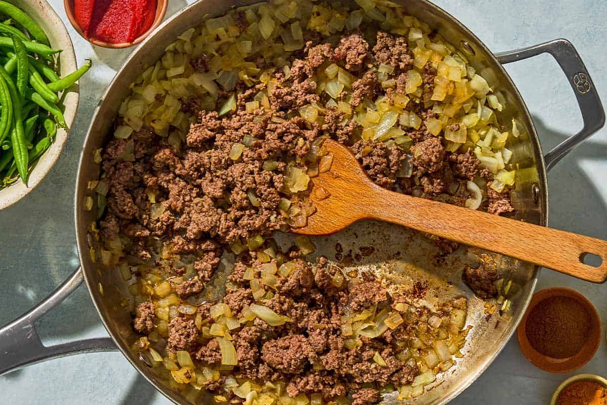 The dicd onion, garlic and ground meat for the loobia polo being cooked in a skillet with a wooden spoon. Next to this is is a bowl with the green beans, and bowls of tomato paste, turmeric and ground cinnamon.