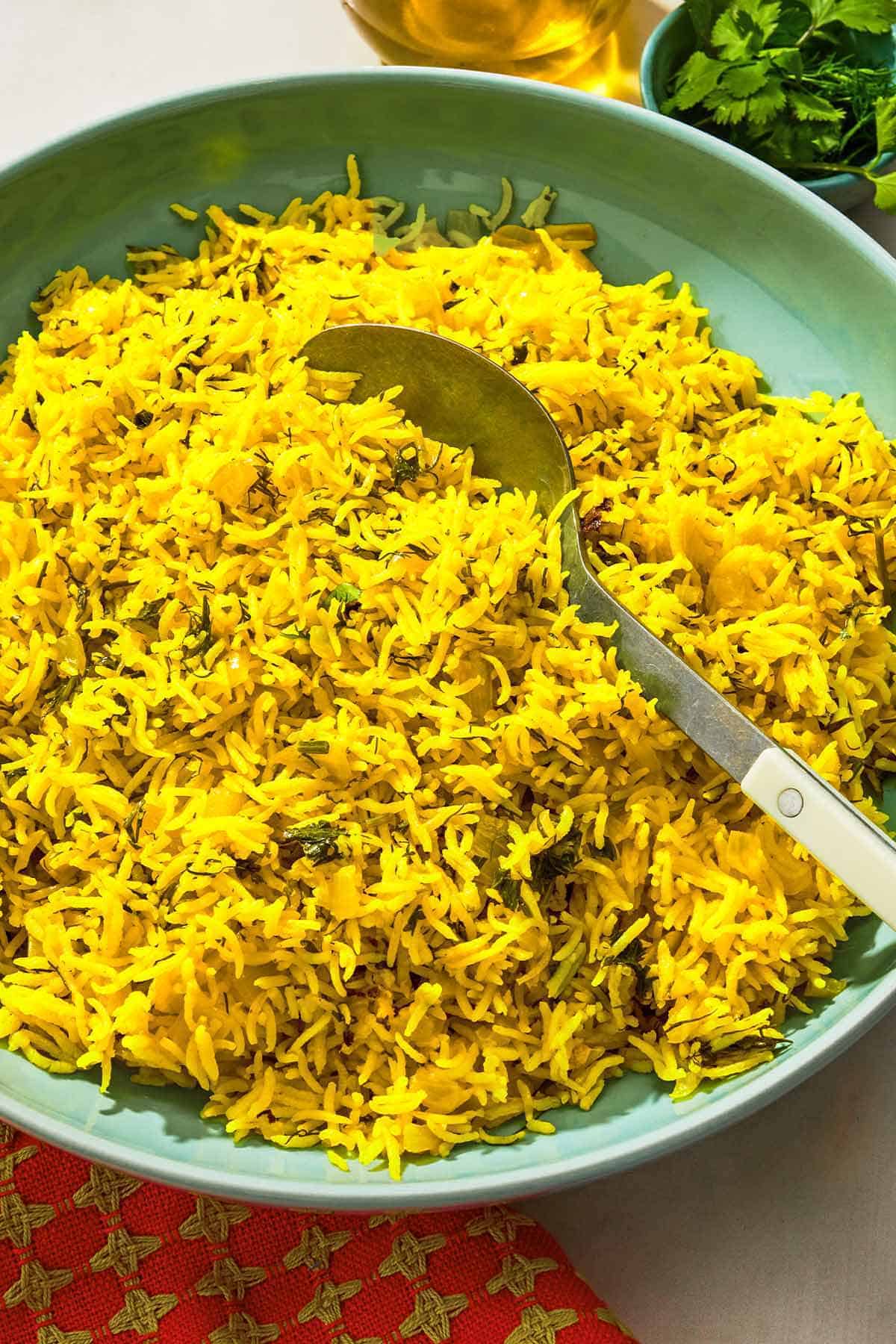 Turmeric rice in a serving bowl with a spoon next to a bowl of cilantro and a bottle of olive oil.