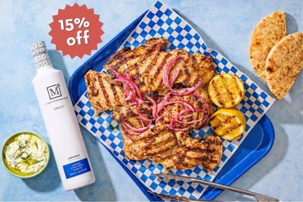 Grilled chicken thighs on a platter with a bottle of The Mediterranean Dish olive oil and a 15% off sales badge.