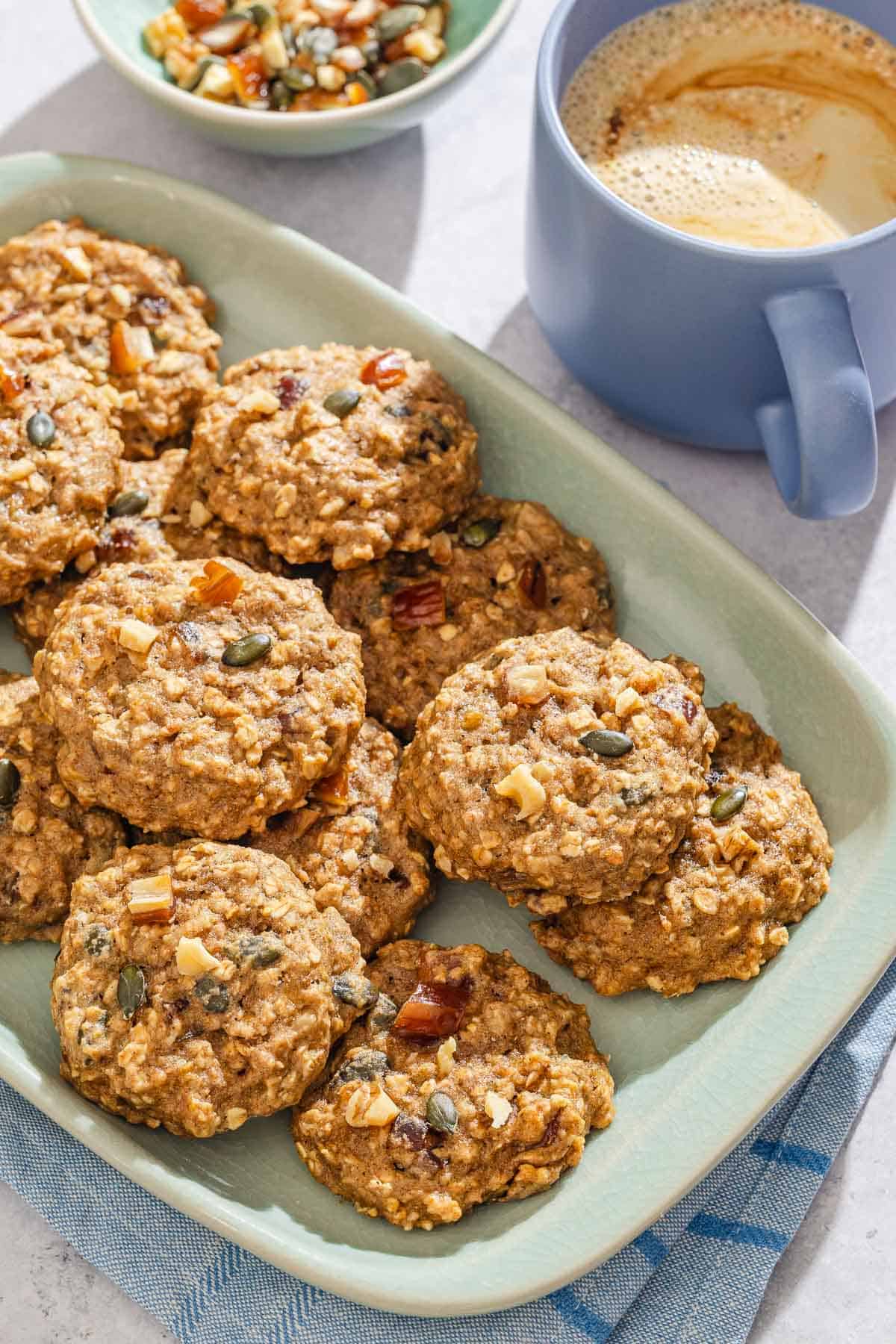 Breakfast cookies on a platter next to a bowl of nuts, seeds and dried fruit and a cup of coffee.
