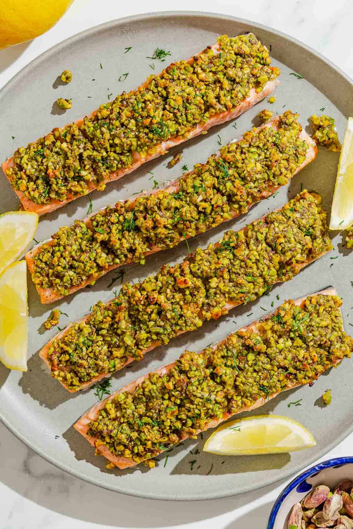 Four pistachio crusted salmon fillets on a platter with lemon wedges.