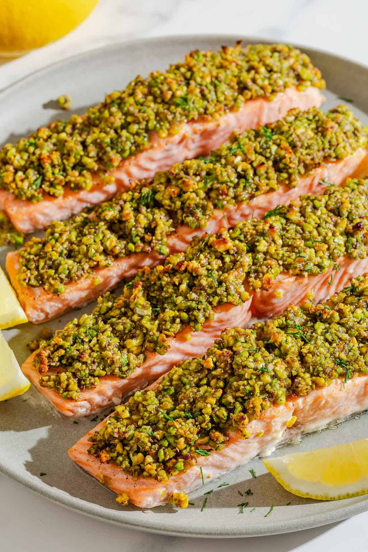 Four pistachio crusted salmon fillets on a platter with lemon wedges.