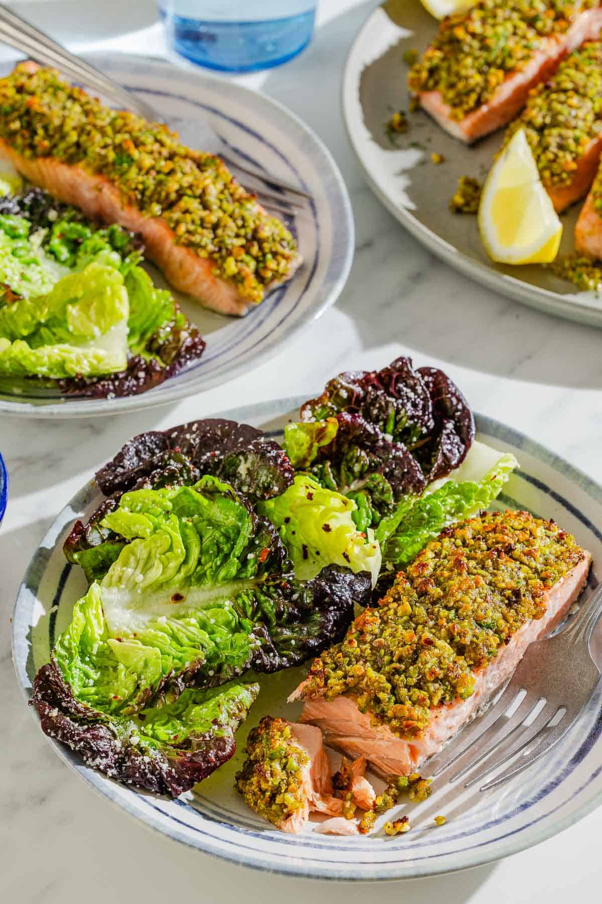Two servings of the pistachio crusted salmon fillet on plates with salads and forks. Next to these is a platter with the rest of the salmon and a lemon wedge.