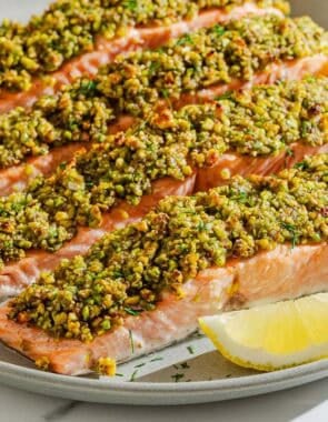 Four pistachio crusted salmon fillets on a platter with lemon wedges.