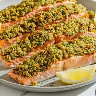 Four pistachio crusted salmon fillets on a platter with lemon wedges.