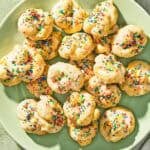 Italian lemon cookies on a platter next to a bowl of non-pareils, a stack of 2 plates, and a lemon.